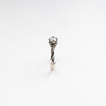 This exquisite solitaire ring features a 2.5mm wide band, intricately hand-carved from luxurious 18ct white gold. The band is designed with a delicate vine motif, symbolising the intertwining of natural beauty and sophisticated craftsmanship.
